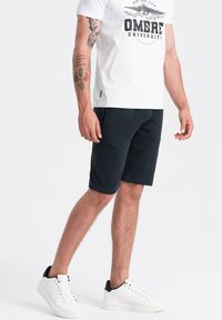Shorts bleu marine en tissu doux, dotés d'une taille élastique et de poches latérales. Portés avec un t-shirt blanc et des baskets blanches.