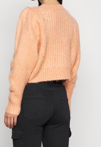 Lätt stickad pullovertröja i ljus persika, med ribbad halsringning och manchett. Korta design som går till mitten av ryggen. Kombinerad med svarta byxor.