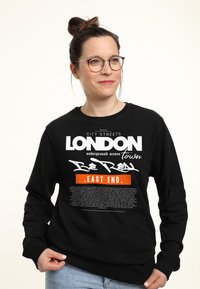 Svart sweatshirt med grafisk textdesign i vitt och orange. Texten inkluderar "LONDON" och "EAST END" med ytterligare tryckta detaljer.