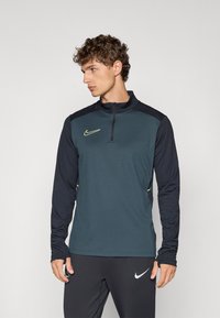 Nike lange mouwen zip-up top in donkerteal en marine, met een slim fit, contrasterende schouderpanelen en reflecterende logo-accenten.