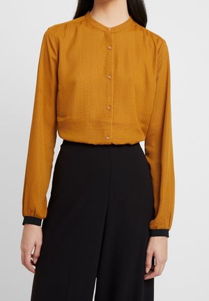 Femme portant un chemisier à manches longues jaune moutarde avec un petit motif à carreaux et des détails de poignets noirs, associé à un pantalon noir taille haute.