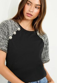 Sort top med korte ærmer, der har et tekstureret sort og hvidt tweed-design, prydet med sølvknapper på skulderen.