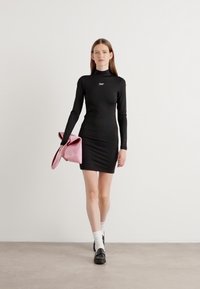 Svart langermet minikjole med en tettsittende silhuett og rund halslinning. Modellen holder en myk rosa veske, kombinert med hvite sokker og svarte sko.