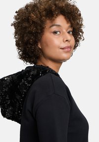 Felpa nera con cappuccio tempestato di paillettes. Il tessuto è morbido e le paillettes offrono un contrasto testurizzato e scintillante lungo il cappuccio.