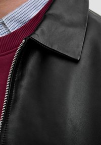 Schwarze Lederjacke mit glatter Textur, ausgestattet mit einem umgeschlagenen Kragen und silbernem Reißverschluss. Darunter ist ein burgunderfarbener, gerippter Pullover sichtbar.