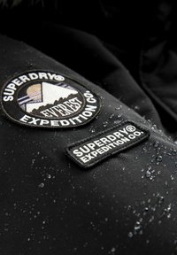 Čierna nepremokavá bunda má na látke dva dekoračné nášivky: kruhové logo a obdĺžnikovú nálepku, pričom obidve jasne zobrazujú "Superdry® Expedition Co."