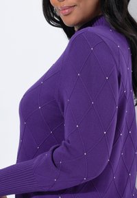 Žena v fialovém svetru s diamantovým prošíváním a malými stříbrnými nýty, ukazující částečný boční profil a rameno.