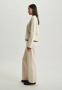 Pull en tricot côtelé crème avec des accents marron, associé à un pantalon large beige clair. Texture lisse, design minimaliste, porté avec des chaussures noires.