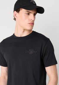 Tricou din bumbac negru cu decolteu rotund și mâneci scurte. Oferă un logo brodat tonal pe piept. Textură netedă, croială casual.