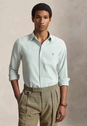 Młody mężczyzna z podwiniętymi rękawami, ubrany w jasnoniebieską koszulę na guziki i brązowe spodnie w kratę, stoi z jedną ręką w kieszeni.