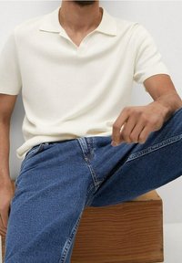 Hombre con una camiseta polo blanca de mangas cortas y jeans azules, sentado sobre una caja de madera frente a un fondo liso.