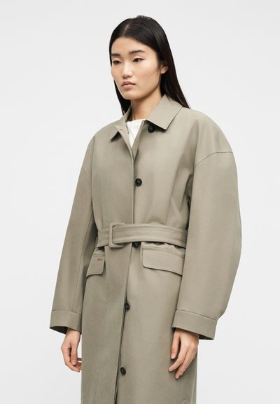 Γυναίκα με μακριά σκούρα μαλλιά που φοράει ένα μπεζ καμπαρντίνα με ζώνη και μαύρα κουμπιά, πάνω σε απλό λευκό φόντο.