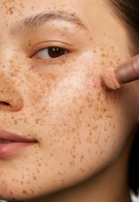 Primo piano di un volto con lentiggini di colore marrone chiaro, texture della pelle liscia e una punta delle dita che applica un prodotto di tonalità pesca utilizzando un applicatore morbido.
