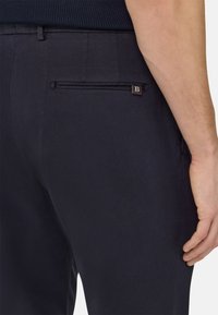 Navy tailored trousers featuring a textured fabric, slim fit, and a back pocket with a metal zip and embossed logo accent.