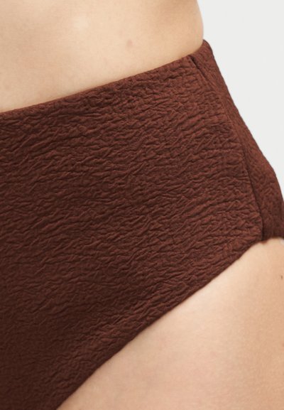 Gros plan sur la taille d'une personne portant une culotte marron texturée taille haute, sur un fond clair uni.