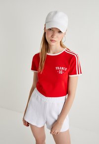 Rode katoenen T-shirt met witte randen en de opdruk "FRANKRIJK 16", gecombineerd met witte katoenen shorts. De outfit heeft een casual, sportieve uitstraling.