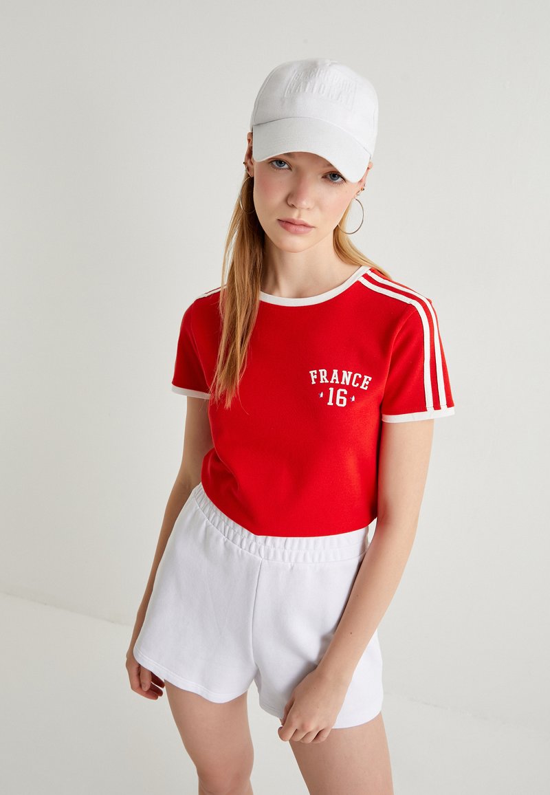 Rode katoenen T-shirt met witte randen en de opdruk "FRANKRIJK 16", gecombineerd met witte katoenen shorts. De outfit heeft een casual, sportieve uitstraling.