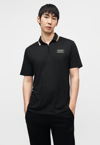 Μαύρο πόλο μπλουζάκι με φερμουάρ στο γιακά, διακοσμημένο με χρυσές ρίγες και κονκάρδα με το λογότυπο "EA7" στο αριστερό στήθος. Κοντά μανίκια και απαλό ύφασμα.