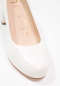 Escarpin en cuir blanc avec bout arrondi, talon épais et texture lisse. Intérieur avec logo doré. Semelle de teinte bois clair.