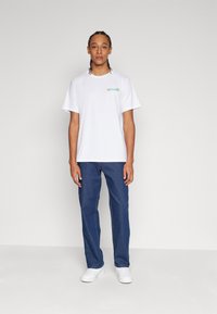 Camiseta de algodón blanca con mangas cortas, con un gráfico multicolor en el pecho, combinada con jeans de denim azul y zapatillas blancas.