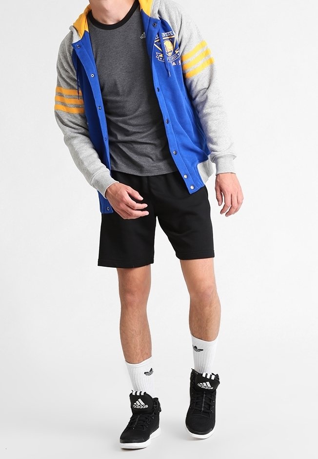 Chaqueta varsity azul y gris con rayas amarillas, pantalones cortos negros, zapatillas negras de caña alta y calcetines blancos; aspecto atlético casual.