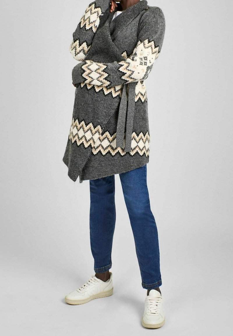 Personne portant un cardigan en maille gris avec des motifs zigzag crème, un jean bleu et des baskets blanches, debout sur un fond uni.