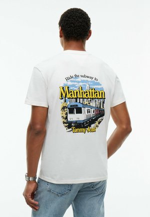 Person trägt weißes T-Shirt mit bunter Grafik einer U-Bahn und der Skyline von Manhattan, Text lautet "Ride the subway to Manhattan Tommy Jeans."