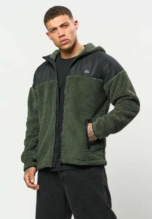 Mann mit kurzen Haaren, der eine grüne und schwarze Fleecejacke und schwarze Hose trägt, steht mit einer Hand in der Tasche und einem seriösen Gesichtsausdruck.