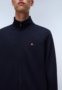 Navy zip-up jacket with high collar, made of textured fabric. Features a small red and white embroidered flag logo on the chest.