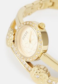 Goldfarbene Armbanduhr mit einem runden Ziffernblatt, einem strukturierten hellgoldenen Ziffernblatt und einer Lünette, die mit klaren Kristallen verziert ist. Mit floralen Akzenten am Armband.