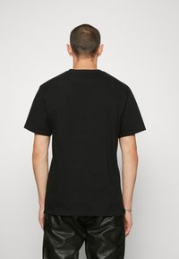 Camiseta de algodón negra de manga corta y cuello redondo, vista por la parte de atrás, combinada con pantalones negros texturizados. Diseño simple sin gráficos.