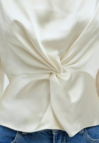 Crème-kleurige blouse met een gedraaide knoopdetail aan de taille, gladde stof en een licht getailleerde pasvorm, gecombineerd met blauwe denim jeans.