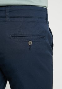 Detailní záběr na zadní kapsu tmavě modrých kalhot s knoflíkovým zapínáním a viditelným šitím na pase a v oblasti kapsy.