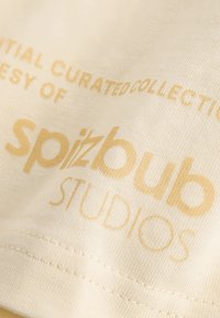 Cream cotton fabric with gold text, featuring "spizbub STUDIOS" and "INITIAL CURATED COLLECTION," with fine stitching along the edge.