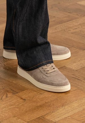 Beige suède sneakers met witte zolen gedragen bij donkere jeans, staand op een bruine houten parketvloer.