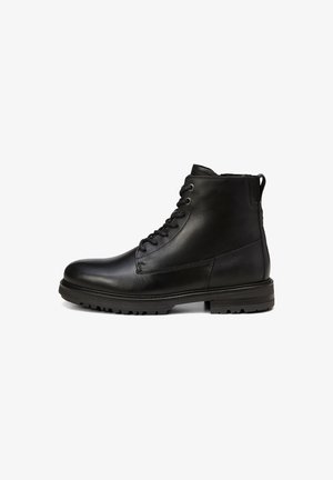 Bottes chelsea en cuir noir avec une forme arrondie à l'avant, un laçage sur le devant et une semelle texturée. Elles disposent d'une languette à l'arrière et de détails de couture minimalistes.