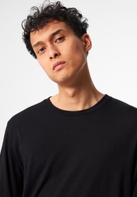 Schwarzes Langarmshirt mit rundem Halsausschnitt. Glatte Textur, minimierte Nähte und figurbetonte Passform. Das Modell präsentiert das Shirt vor einem neutralen Hintergrund.