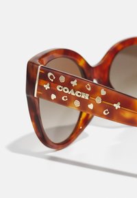 Ochelari de soare cu montură țestoasă, lentile maro, cu logo-ul auriu "COACH" și motive decorative pe brațe, design rotund, textură lucioasă.