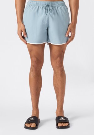 Hombre con pantalones cortos de baño azul claro con ribete blanco y sandalias deslizantes negras de Champion, de pie con las manos relajadas a los lados.