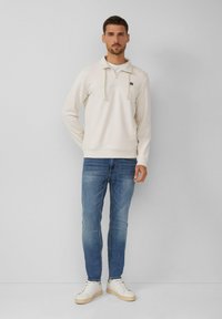 Sudadera color crema con media cremallera, que incluye un bolsillo canguro y capucha con cordón, combinada con jeans azules y zapatillas blancas.