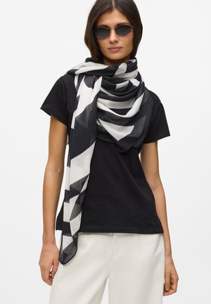 Žena nosící sluneční brýle, černé tričko, bílé kalhoty a velký černobílý šátek s geometrickým vzorem přehozený přes krk.