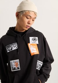 Sudadera negra con varios parches gráficos, incluyendo diseños en naranja y monocromos, cordones en la capucha y una textura de tela suave.