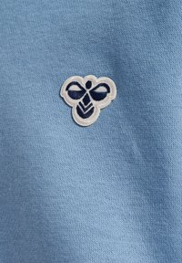 Lichtblauwe stof met een gestructureerde gebreide textuur, met een wit en marineblauw geborduurd bijenlogo aan de linkerkant.