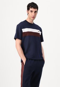 Ναυτικό βαμβακερό μπλουζάκι με οριζόντιες άσπρες και μπορντό ρίγες; διαθέτει ένα μικρό λογότυπο Lacoste στο αριστερό στήθος. Το μοντέλο έχει τα χέρια στις τσέπες.