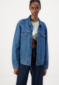 Blå denimjakke med et intrikat paisley-mønster, knappet front, to brystlommer og en avslappet passform. Brukt over en lys grønn topp.