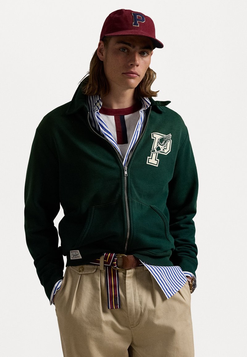 Felpa verde con zip, con un emblema, indossata sopra una camicia a righe blu e bianca, pantaloni beige e una cintura multicolore.