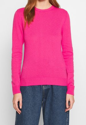 Kvinde iført en knaldrød lyserød langærmet sweater og løstsiddende blå jeans, stående mod en helt hvid baggrund.