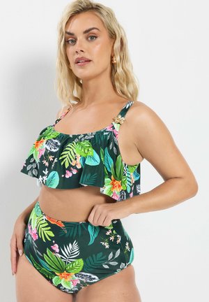 Blonde vrouw die een groen tropisch bloemenbikinitwee-delig met gerimpelde top en hoog getailleerde broekjes draagt, staand tegen een witte achtergrond.