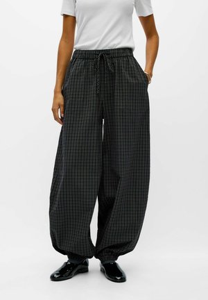 Modèle portant un pantalon ample à carreaux foncés, avec une taille à cordon de serrage et des chevilles froncées, associé à un T-shirt blanc et à des chaussures noires.