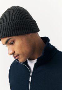 Dark gray knitted beanie with ribbed texture, worn with a navy zip-up jacket over a white shirt, displaying a close-up profile view.